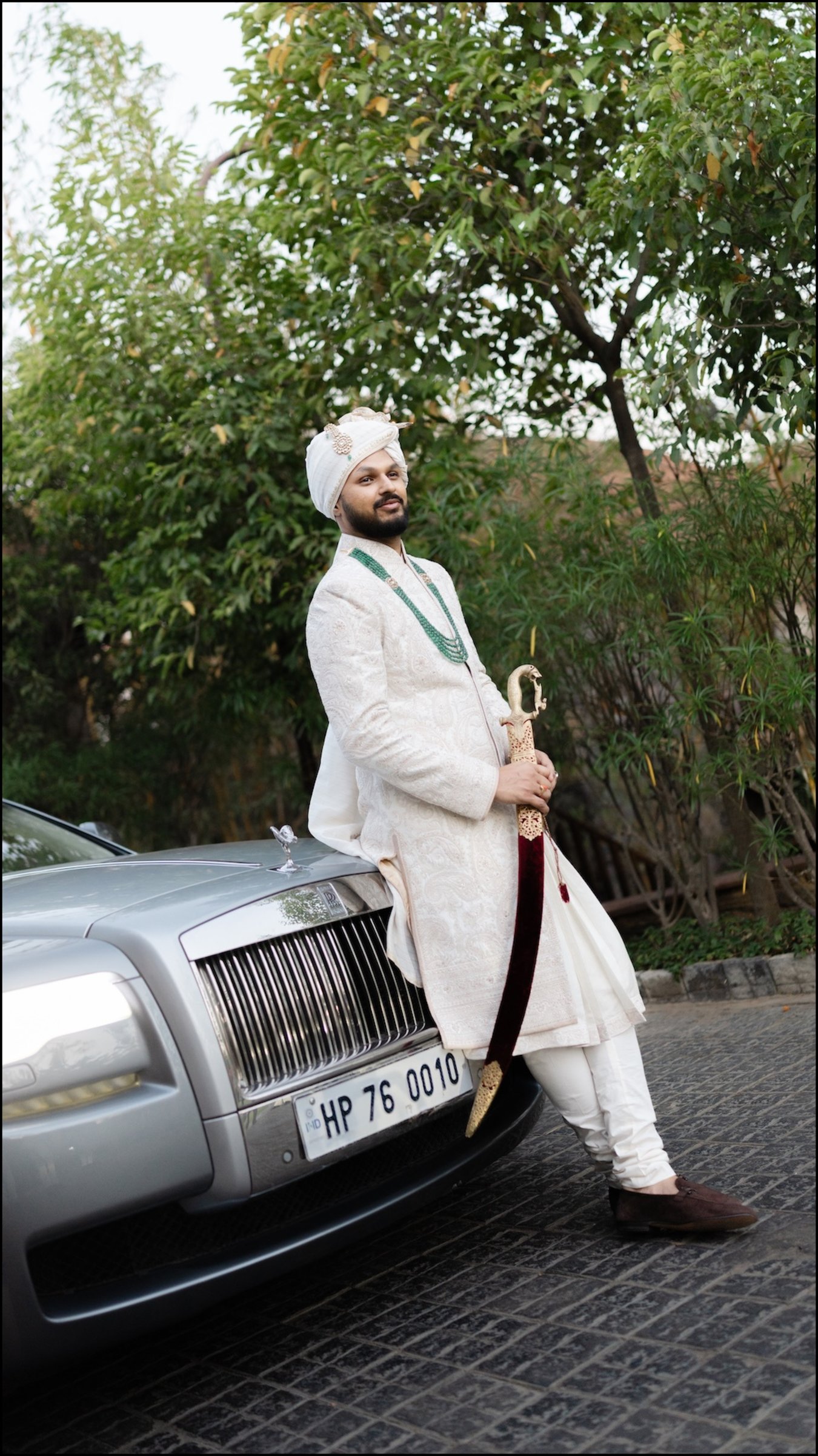 Groom in an ivory sherwani holding a sword beside a silver luxury wedding car